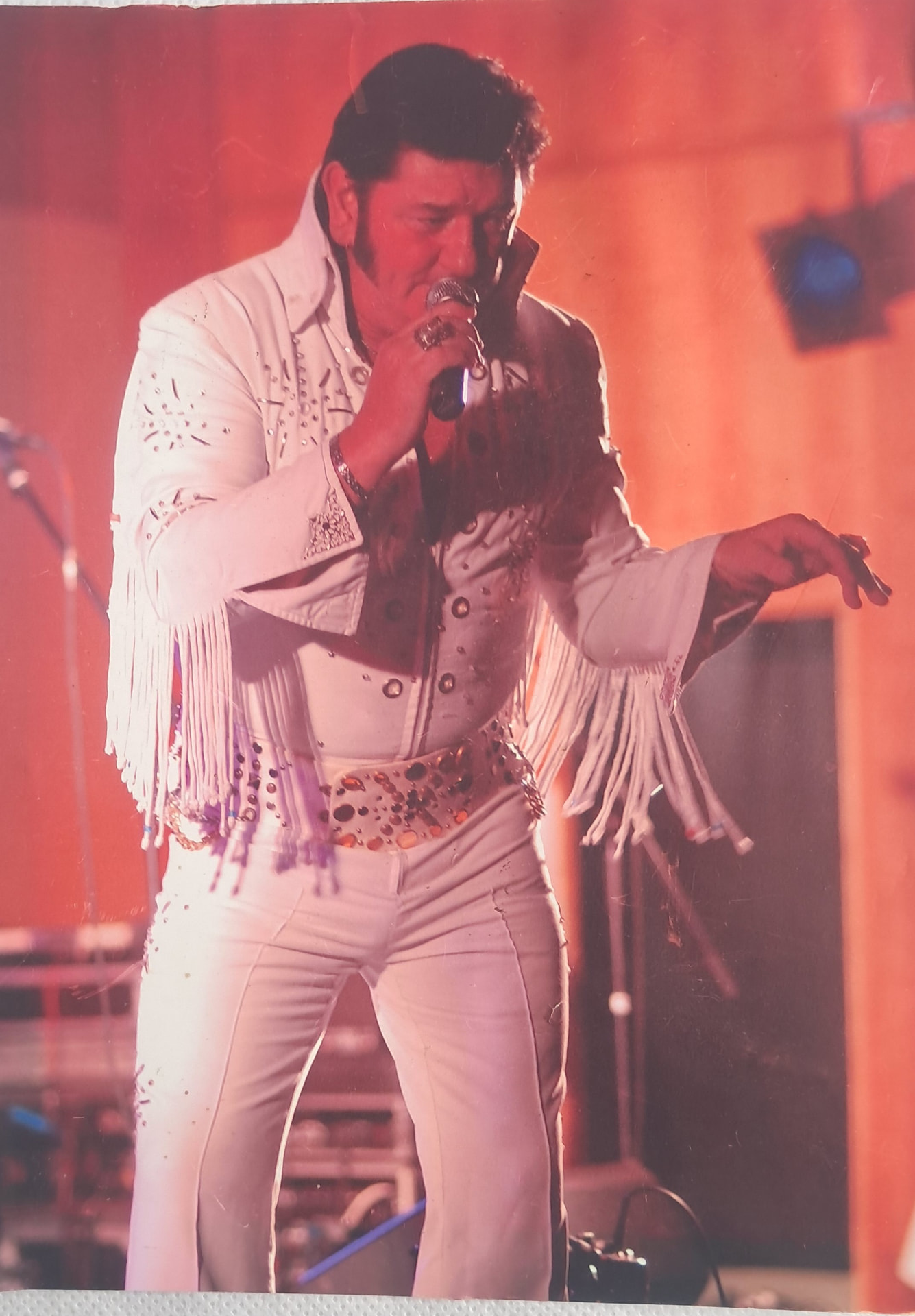 Greg Traynor performing as an Elvis tribute artist on stage in a white jumpsuit, holding a microphone